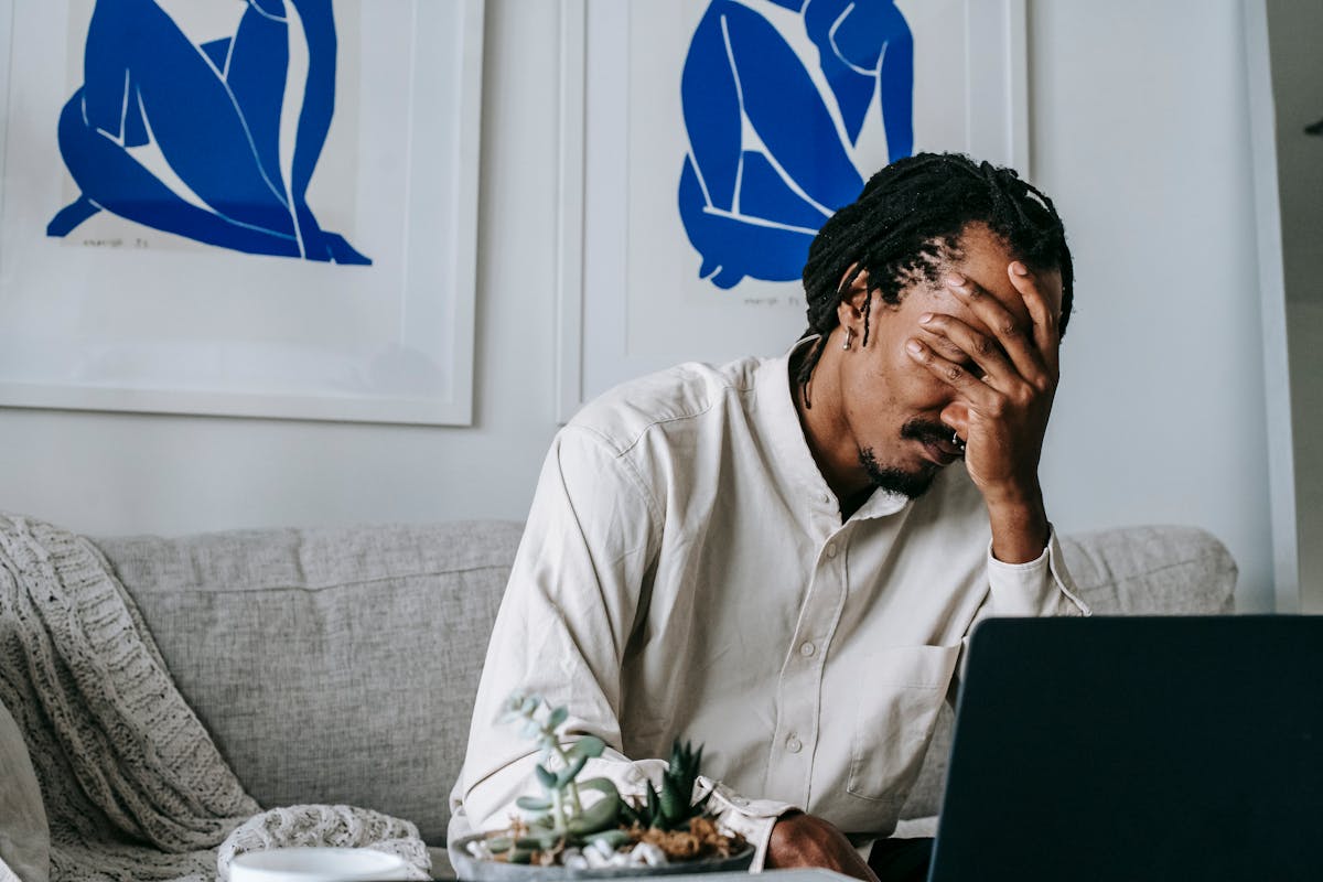 Upset young black guy covering face with hand while working remotely on netbook